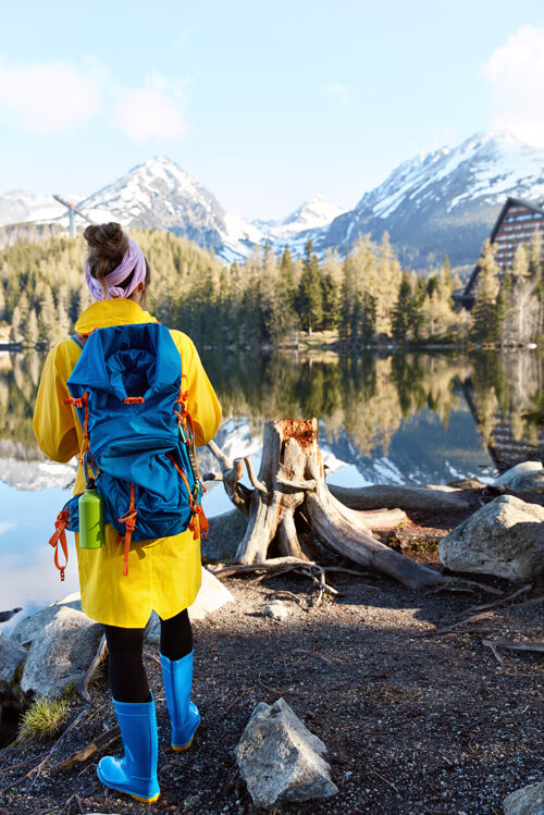 姿势背着背包的游客站在山河边 欣赏风景秀丽的大自然 穿着黄色的长袍靴子女性探索