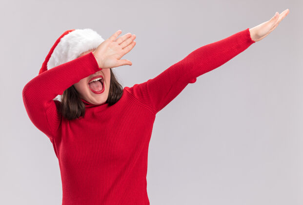 站着有趣的年轻女孩穿着红色毛衣 戴着圣诞帽 站在白色的背景上 开心而兴奋地打着手势圣诞老人毛衣女孩
