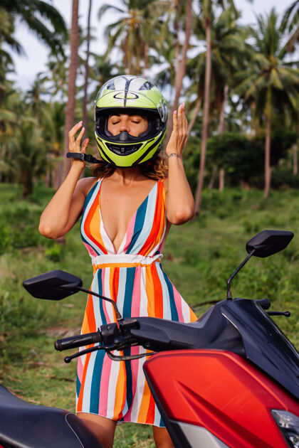 户外在棕榈树下热带田野的丛林中 穿着黄绿色摩托车头盔和彩色浅夏装的美丽骑手妇女的肖像幸福人手掌