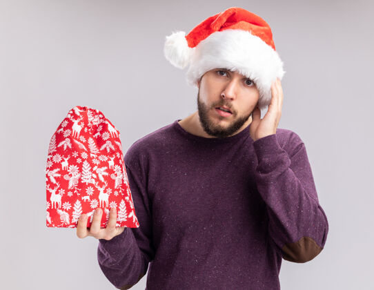 焦虑年轻人穿着紫色毛衣 戴着圣诞帽 手里拿着红包 手里拿着礼物 困惑地看着镜头 非常焦急地站在白色的背景下抱站困惑