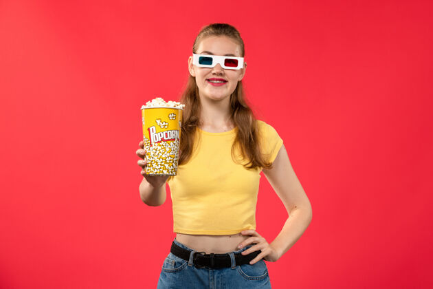 年轻女性前视图年轻女性手持爆米花包 戴着d型太阳镜 站在浅红墙上的电影院里护目镜爆米花电影