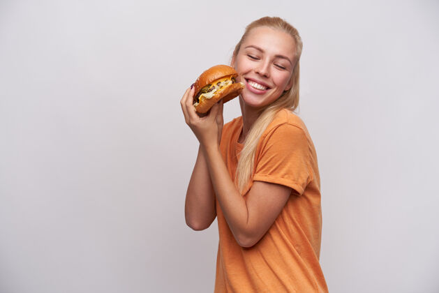 T恤快乐的年轻长发金发女人 随意的发型 手里拿着美味的新鲜汉堡包 闭着眼睛 站在白色的背景下 欢快地笑着摆姿势休闲情绪