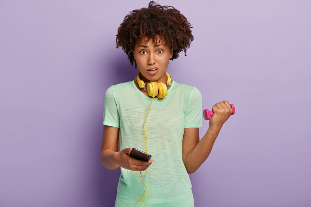 非洲迷惑不解的黑皮肤女子手持智能手机 从手机播放列表中选择健身曲目T恤运动健康