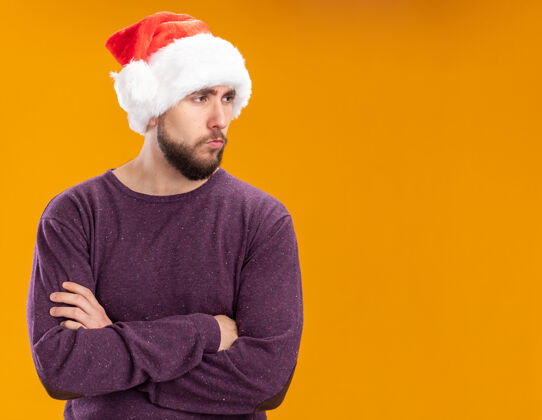 圣诞老人一个穿着紫色毛衣 戴着圣诞帽的年轻人站在橙色的背景下 严肃的脸朝旁边望去帽子年轻站着