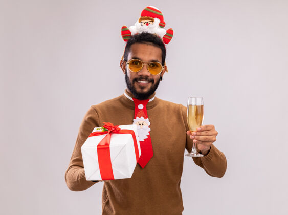 礼物快乐的非洲裔美国人 身穿棕色毛衣 头戴圣诞老人戒指 打着有趣的红色领带 手里拿着礼物 手里拿着一杯香槟 站在白色背景下 面带微笑地看着摄像机香槟毛衣非洲