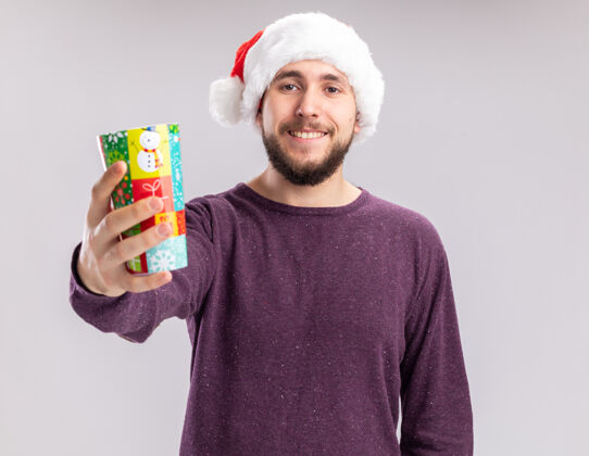 毛衣穿着紫色毛衣 戴着圣诞帽的年轻人展示着五颜六色的纸杯 站在白色背景下 面带微笑地看着相机帽子圣诞老人脸