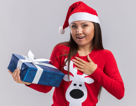 放着笑容满面的亚洲小女孩戴着圣诞帽拿着毛衣拿着礼盒手放在白色背景上孤立的自己身上女孩微笑手