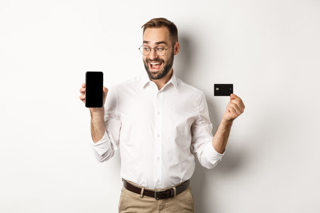 信用商务和网上支付笑容可掬的帅哥展示手机屏幕和信用卡 站着员工时髦胡子