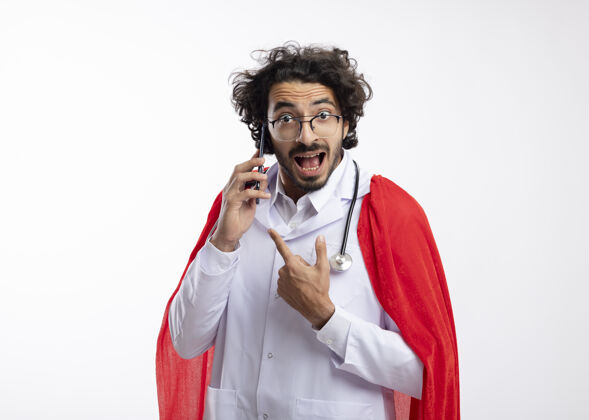 听诊器兴奋的年轻白种人超级英雄戴着眼镜 身穿医生制服 披着红色斗篷 脖子上戴着听诊器 指着隔离在白墙上的电话指向男人电话