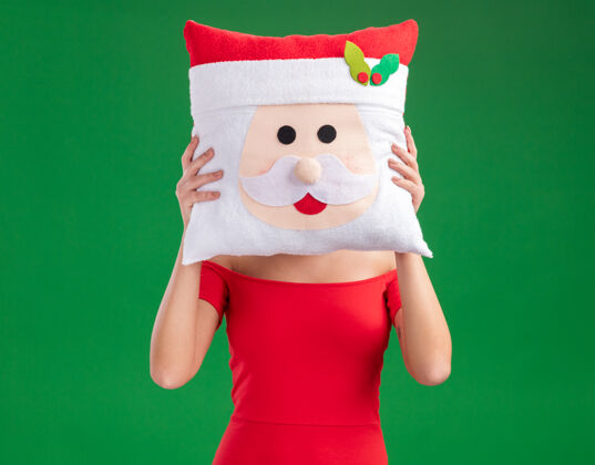 年轻人年轻女孩戴着圣诞帽抱着圣诞老人的枕头 脸上用它隔离着绿色的背景掩护抱着脸