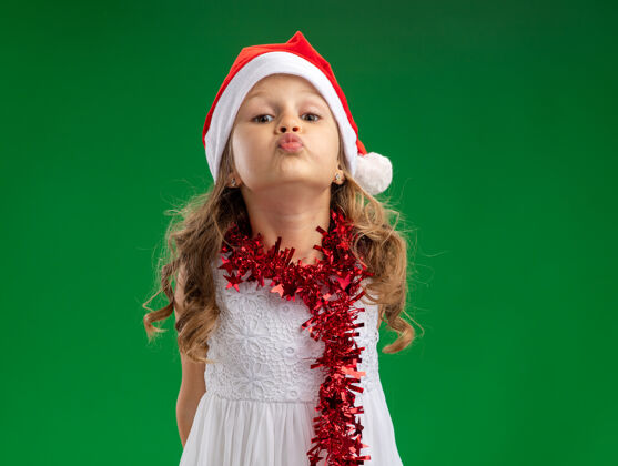 吻可爱的小女孩戴着圣诞帽 脖子上戴着花环 在绿色的背景上显示出与世隔绝的亲吻手势小女孩帽子