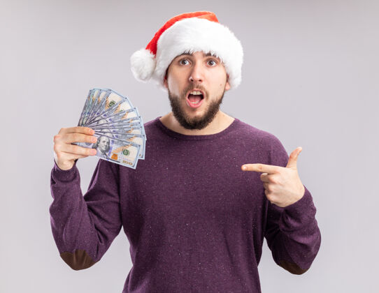 索引一个穿着紫色毛衣 戴着圣诞帽的年轻人拿着现金 用食指指着钞票 站在白色的背景下显得很惊讶毛衣帽子钱