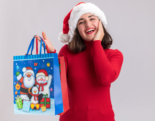 女孩穿着红色毛衣 戴着圣诞帽的小女孩手里拿着五颜六色的纸袋 手里拿着圣诞礼物 站在白色的背景上 满脸幸福地看着相机礼物年轻举行