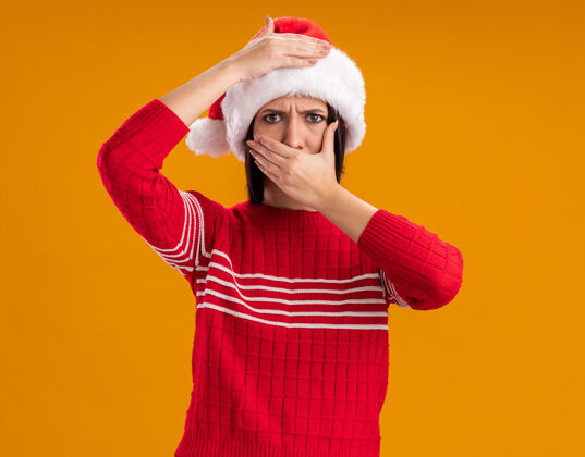 女孩愁眉不展的年轻女孩戴着圣诞帽看着摄像机手放在头上捂住嘴 手孤立在橙色背景上手圣诞老人盖