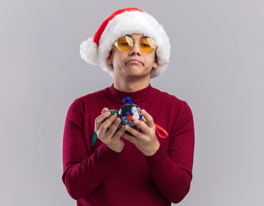 圣诞节戴着圣诞帽戴着眼镜拿着圣诞树球的悲伤的年轻人被隔离在白色背景上眼镜小伙子圣诞树