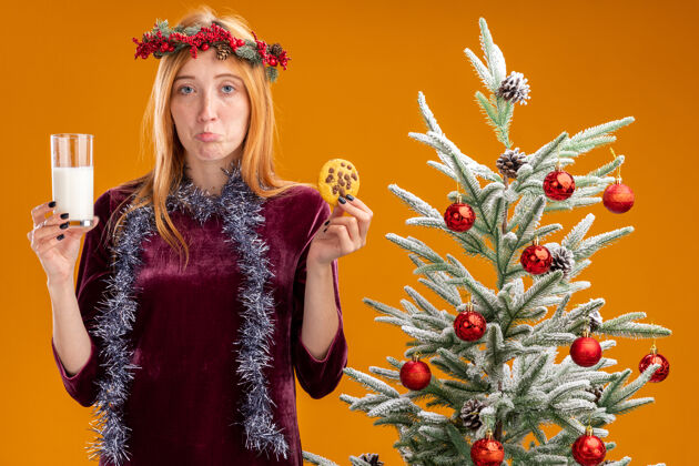 女孩站在圣诞树旁的悲伤的年轻漂亮的女孩 穿着红色的裙子 脖子上戴着花环 手里拿着一杯牛奶和饼干 背景是橙色的饼干脖子牛奶