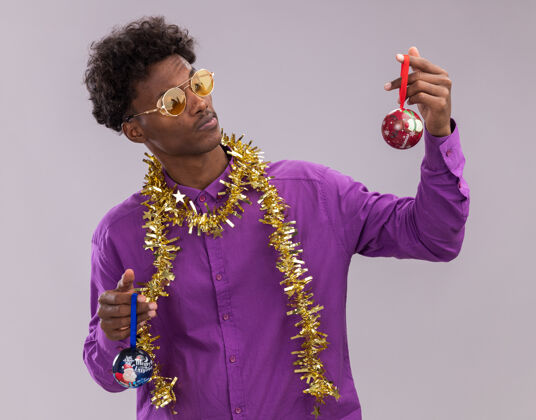 周围一个可疑的美国黑人年轻人戴着眼镜 脖子上戴着金箔花环 手里拿着圣诞饰品 看着其中一个孤立在白色背景上的圣诞饰品眼镜脖子花环