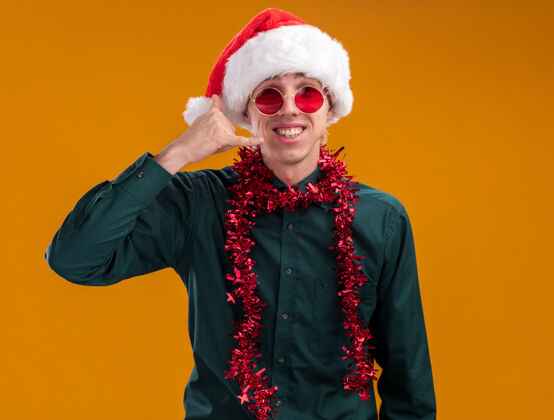 圣诞老人微笑着的年轻金发男子戴着圣诞帽 戴着眼镜 脖子上戴着金箔花环 看着摄像机在橙色背景上做呼号眼镜帽子金发