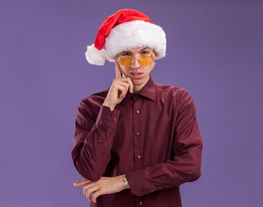 圣诞老人体贴的年轻金发男子戴着圣诞帽 戴着眼镜 在紫色背景下看着摄像机做着孤立的思考动作金发手势眼镜