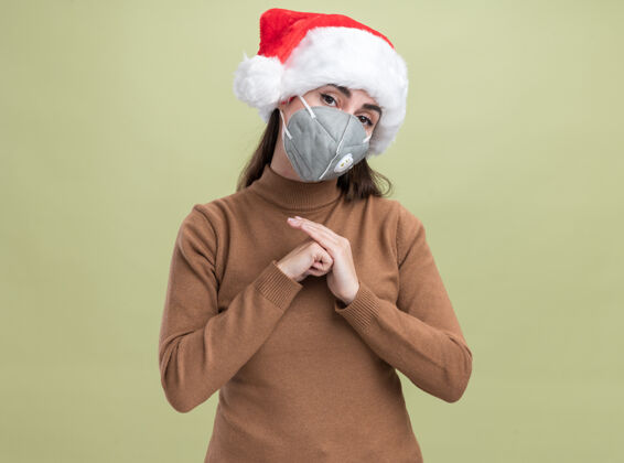 面具高兴的歪着头的年轻漂亮女孩戴着圣诞帽带着医用口罩手牵着手隔离在橄榄绿的背景上高兴倾斜橄榄