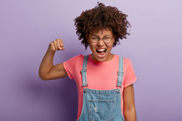 萨拉凡情绪激动的非裔美国女孩举起拳头 从积极的情绪中惊呼 笑脸 穿着粉色t恤和牛仔萨拉凡 站在紫色的墙上我们一起做了 祝贺凝视面部惊呼