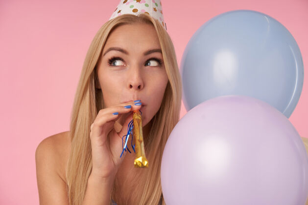 发型特写镜头中 一位喜气洋洋的金发碧眼的长发女士拿着五颜六色的气球在粉色背景上摆姿势 积极地向一边看 嘴里叼着节日派对的号角年轻20多岁迷人