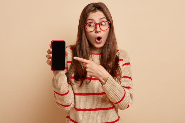 室内惊讶的欧洲年轻女子从惊奇中张嘴 指着手机以空白屏幕为内容模板 设计 身穿米黄色红色条纹套头衫 隔着棕色的墙壁个性屏幕在线