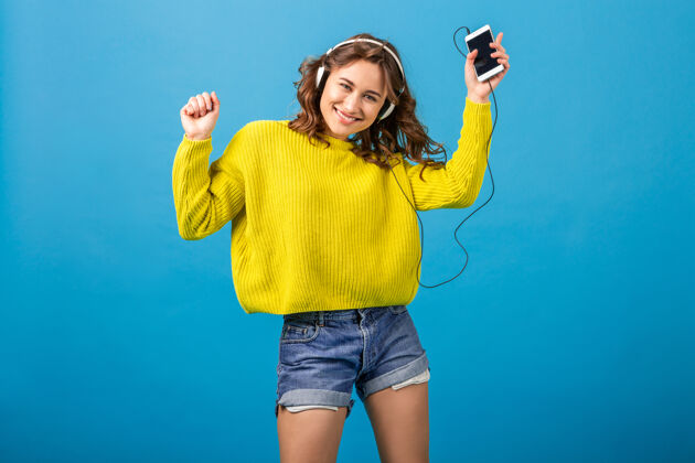 跳跃迷人的笑容可掬的快乐女人戴着耳机跳舞听音乐 穿着时髦的服装 隔离在蓝色的工作室背景下 穿着短裤和黄色毛衣听五颜六色蓝色