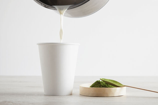 桌子侧视图：木制桌上的白色纸杯和优质有机日本抹茶 为现代拿铁咖啡的制作做好准备演示第三步将一点热牛奶倒进杯子里木材饮料抹茶