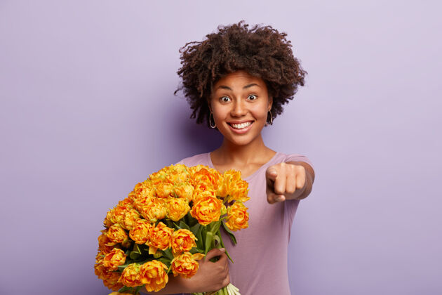 牙关漂亮乐观的女人用食指直指你 邀请你来度假 捧着黄花 穿着休闲t恤 隔着紫色的墙壁 露出愉快的笑容单色乐观积极高兴