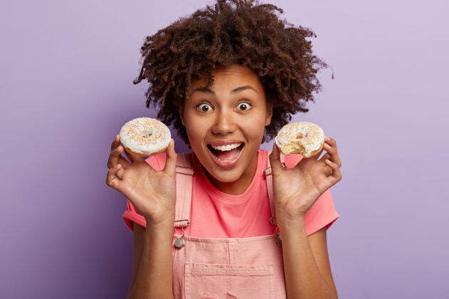 面包店快乐的非洲裔美国妇女有美味的糕点早餐 拿着两个甜甜圈 享受美味的甜点 吃不健康的食物 与紫色墙壁隔离女性甜食美味Pursing卷发