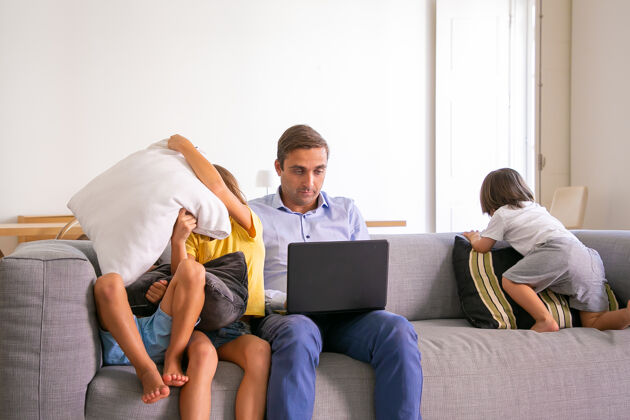 女儿中年爸爸坐在沙发上用笔记本电脑工作 孩子们在他身边玩耍白人爸爸用电脑 孩子们在房间里的教练上玩得很开心数字技术和父亲的概念房间成人孩子
