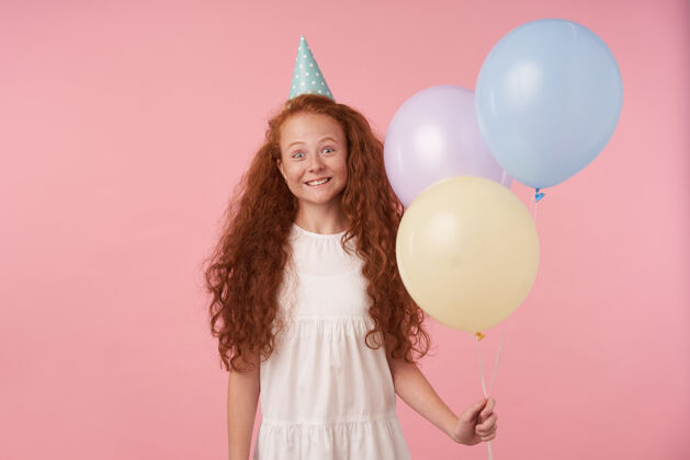 卷曲快乐的红发女孩 长卷发 穿着节日服装 戴着生日帽 在粉色背景下摆姿势 手里拿着气球 表达着真正的积极情绪空气红发肖像