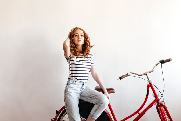 红发穿着条纹t恤坐在自行车上的快乐的姜女玩卷发的快乐的女模特快乐微笑模型