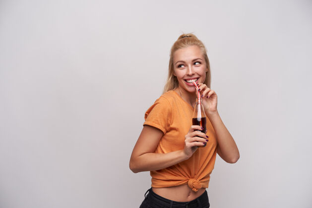 年轻摄影棚照片中 身着橙色t恤的快乐迷人的年轻女子一边喝着稻草汽水 一边迷人地微笑 在白色背景下摆姿势苏打水摆姿势喝酒