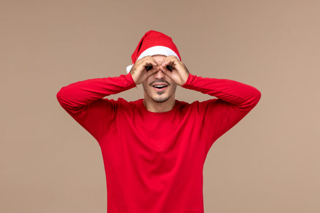 快乐前视图年轻男性透过手指看棕色背景圣诞假期的情绪男性视图看