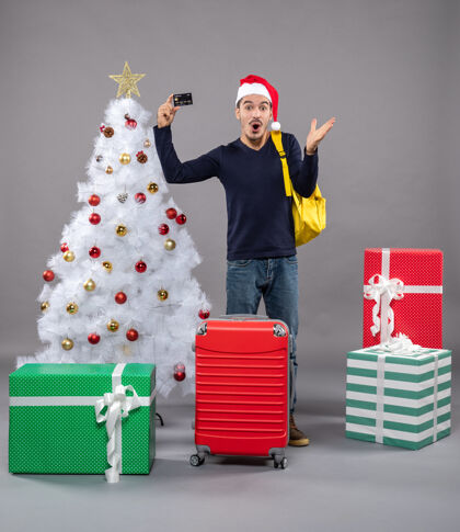 灰色惊讶的年轻人拿着卡片站在圣诞树旁边 礼物是灰色的圣诞老人盒子圣诞