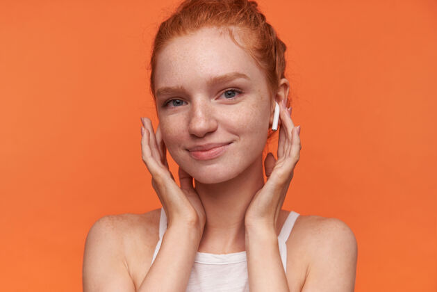 耳机年轻漂亮女人的肖像 戴着狐狸精的发髻 手拿着耳机 对着镜头欢快地微笑 穿着休闲服 隔离在橙色的背景下耳机20多岁头发