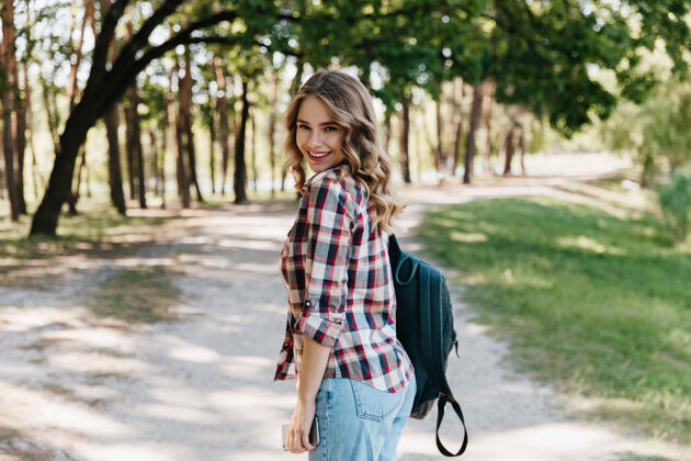 享受穿着格子衬衫和蓝色牛仔裤的快乐女孩站在公园里穿着皮包的女人在春天微笑着旅行休息年轻
