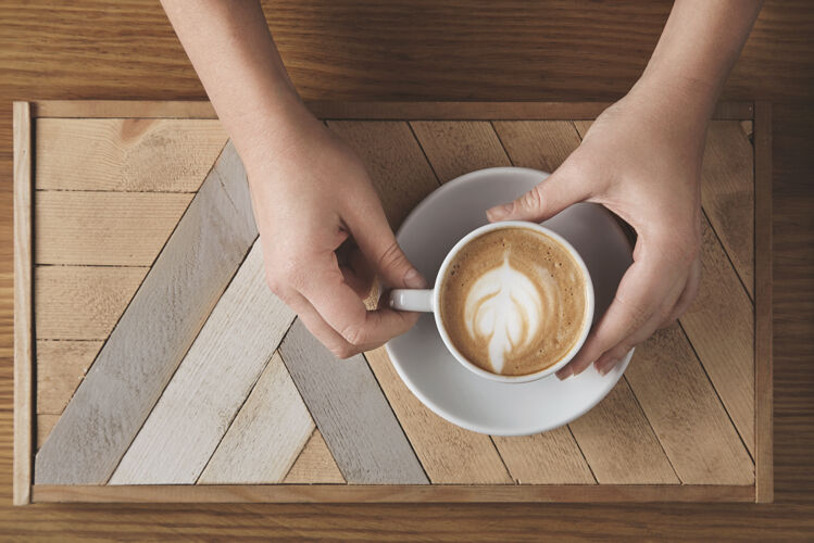 滴美丽的女人手拿着白色的陶瓷 卡布奇诺咖啡 木制的盘子和乡村的桌子树形的牛奶泡沫在上面咖啡店的顶视图销售展示概念咖啡厅咖啡师酒吧