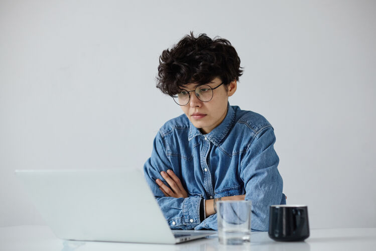 茶一张室内照片 年轻的黑发卷发女子戴着眼镜 手持笔记本电脑 穿着蓝色牛仔裤衬衫 坐在白色背景上 在现代办公室工作时尚自然发型