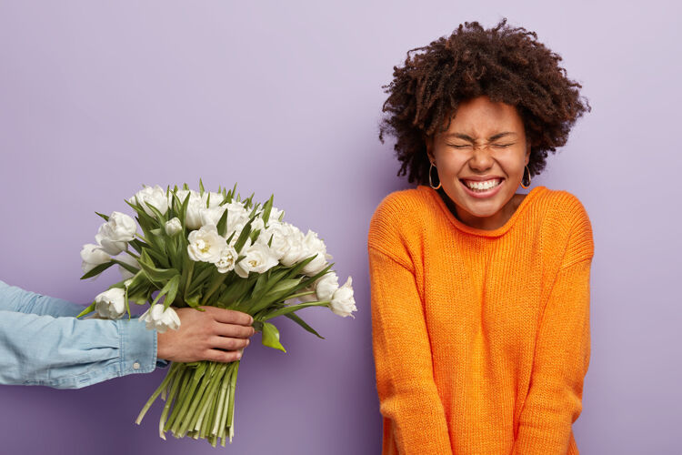 浪漫喜出望外的年轻美国黑人卷发女士在生日当天收到祝贺和鲜花 面目全非的男子伸出双手 赠送春天的白色郁金香 隔离在紫色的墙上大笑女朋友男朋友