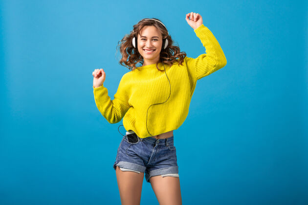 兴奋迷人的笑容可掬的快乐女人戴着耳机跳舞听音乐 穿着时髦的服装 隔离在蓝色的工作室背景下 穿着短裤和黄色毛衣女人耳机音乐