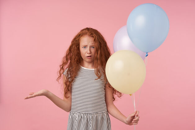 手掌摄影棚照片中 一头红色卷发的小女孩穿着休闲发型 站在粉色背景上 困惑地看着相机 举着气球 抬起手掌未发布情绪化童年