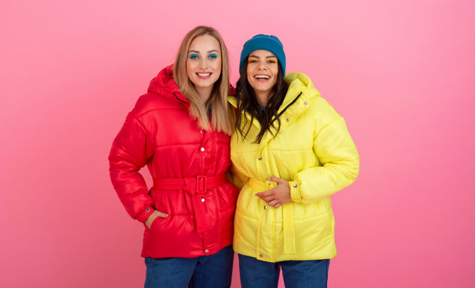 衣服两个迷人的女孩在粉红色的背景下 穿着鲜艳的红色和黄色的彩色冬季羽绒服摆姿势时尚衣服外套