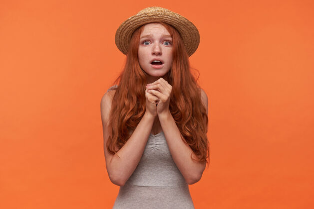 波浪一幅绝望的年轻红发孤独女性的肖像 身着灰色衬衫 头戴水手帽 双手举在一起看着相机 希望能解决她的问题 孤立在橙色背景下20多岁头发衬衫