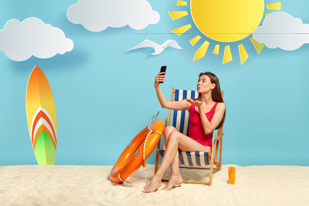 休息可爱的女模特对着智能手机相机吹空中飞吻 自拍 在沙滩椅上摆姿势 穿着红色比基尼沙滩苗条水平
