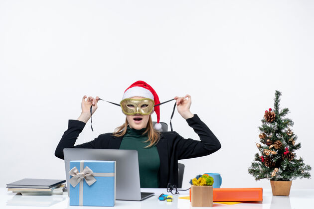 桌子带着圣诞老人帽子戴着面具坐在白色背景的桌子旁的情绪激动的年轻女子面具圣诞老人电脑