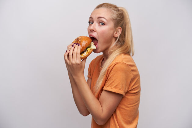 汉堡包摄影棚拍摄的一张照片 一位穿着休闲服的年轻金发美女 手拿美味的汉堡包 站在白色背景下准备吃汉堡包情绪饥饿饮食