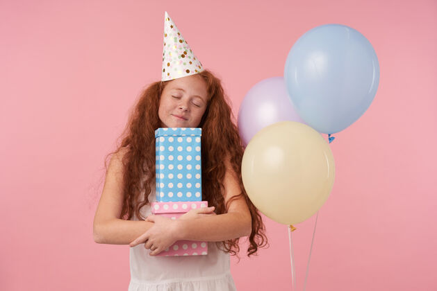 气球积极的红头发卷曲的女孩子在粉红色的工作室背景上摆着彩色气球 抱着礼品盒闭上眼睛 开心地微笑着 穿着节日的衣服和生日帽情绪化高兴庆祝